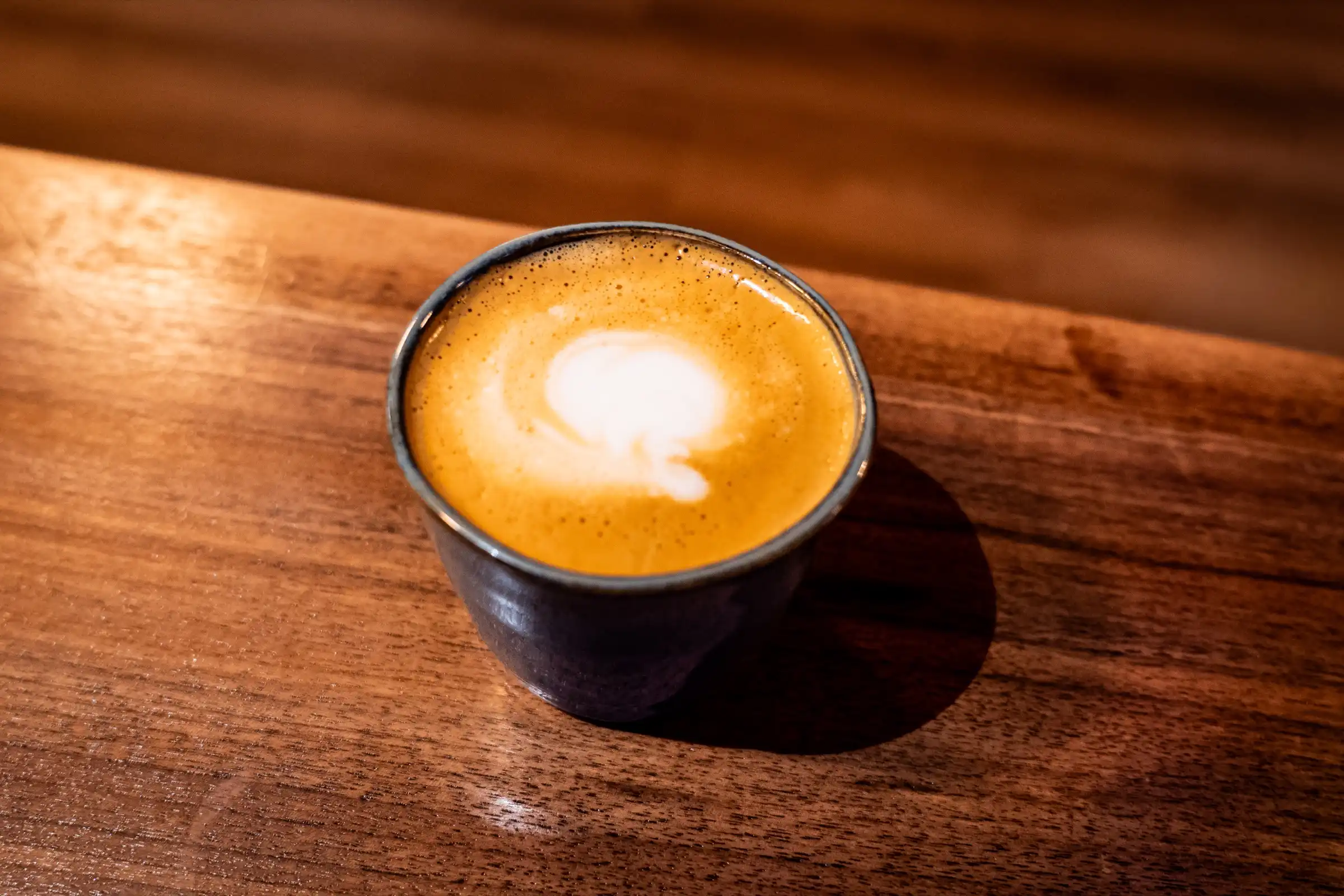 A cortado with a small white heart of foam, in a dark ceramic cup on a walnut bar top.